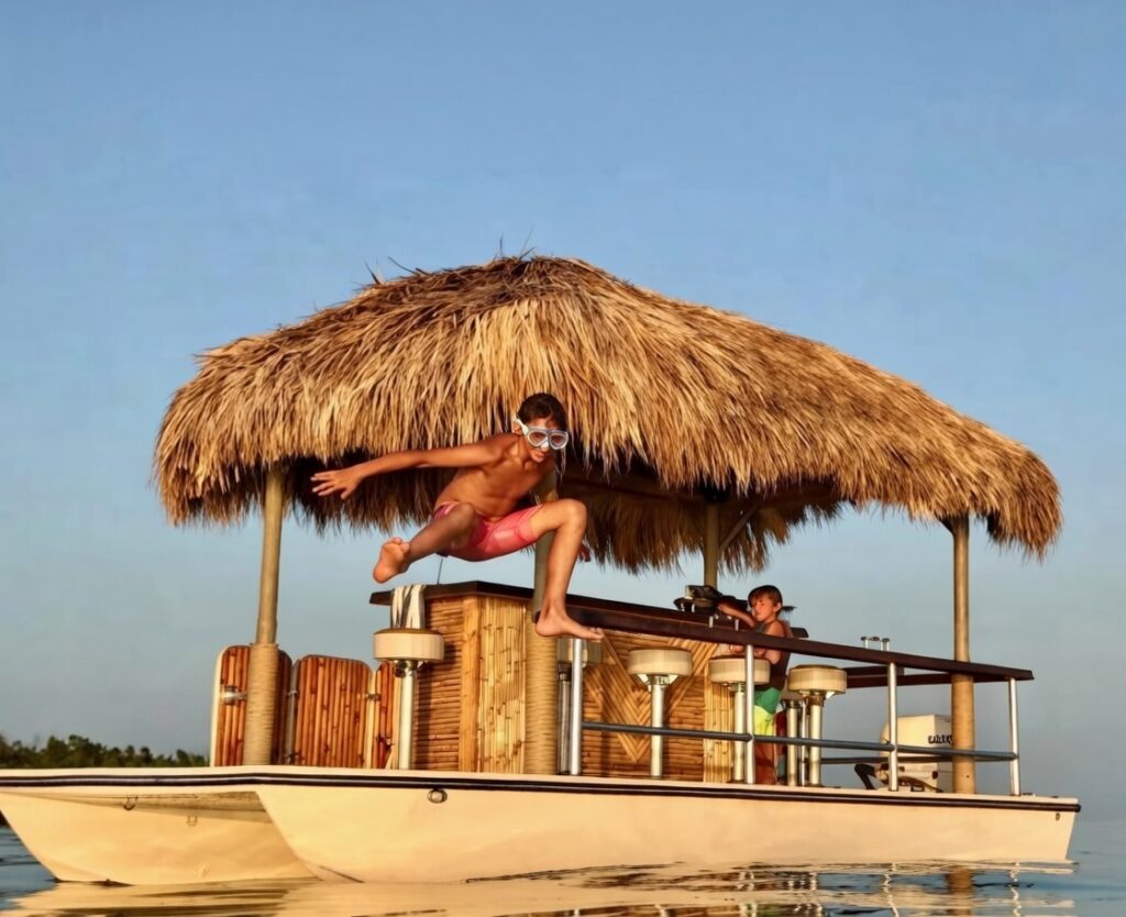 Young boy in snorkel mask jumping off a thatched tiki hut floating pontoon boat into the water during a beautiful sunset in Florida