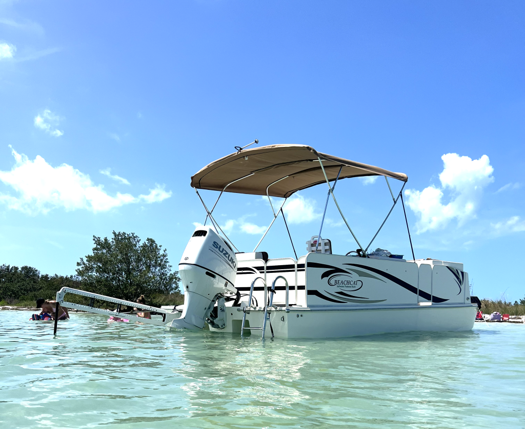 White Beachcat recreational pontoon boat with beige canopy and Suzuki outboard motor floating in clear turquoise saltwater near a sandy beach lined with palm trees along the Texas Gulf Coast